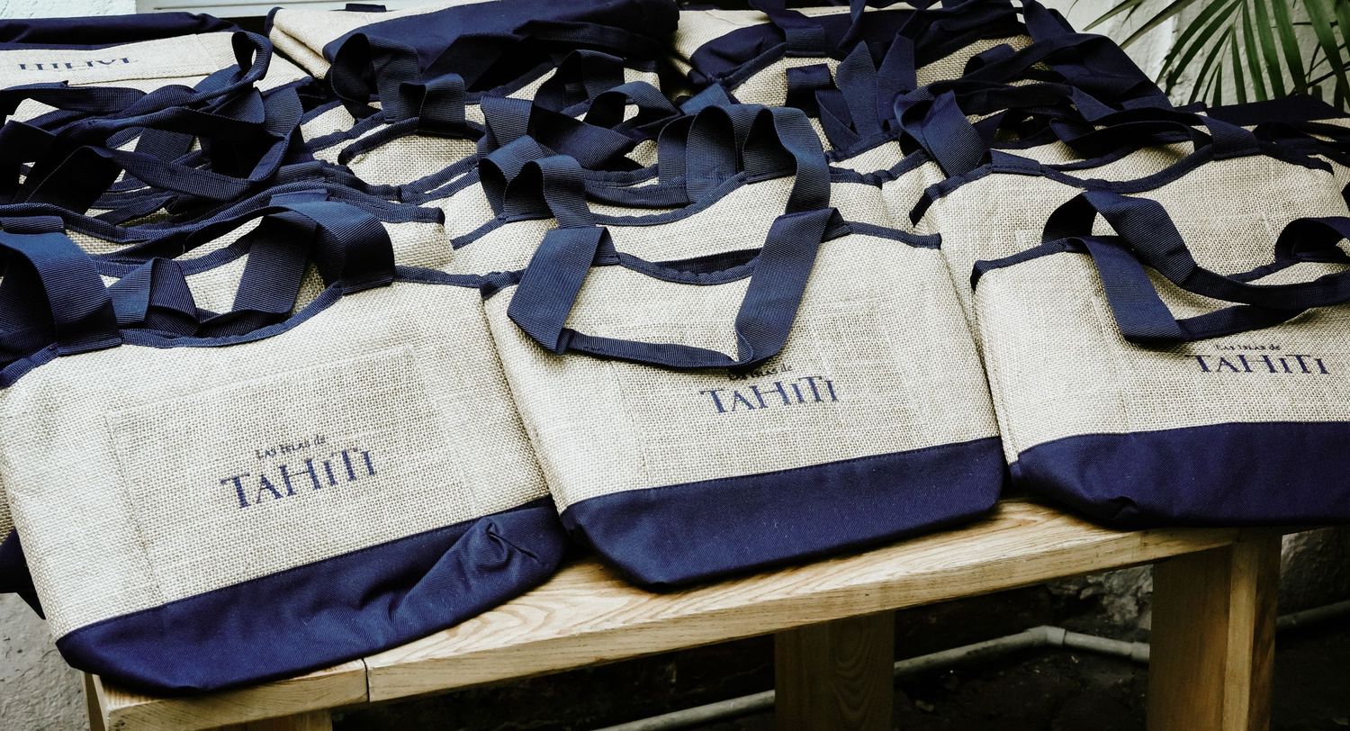 Multiple branded Las Islas de Tahiti jute tote bags with navy blue handles displayed on wooden surface.
