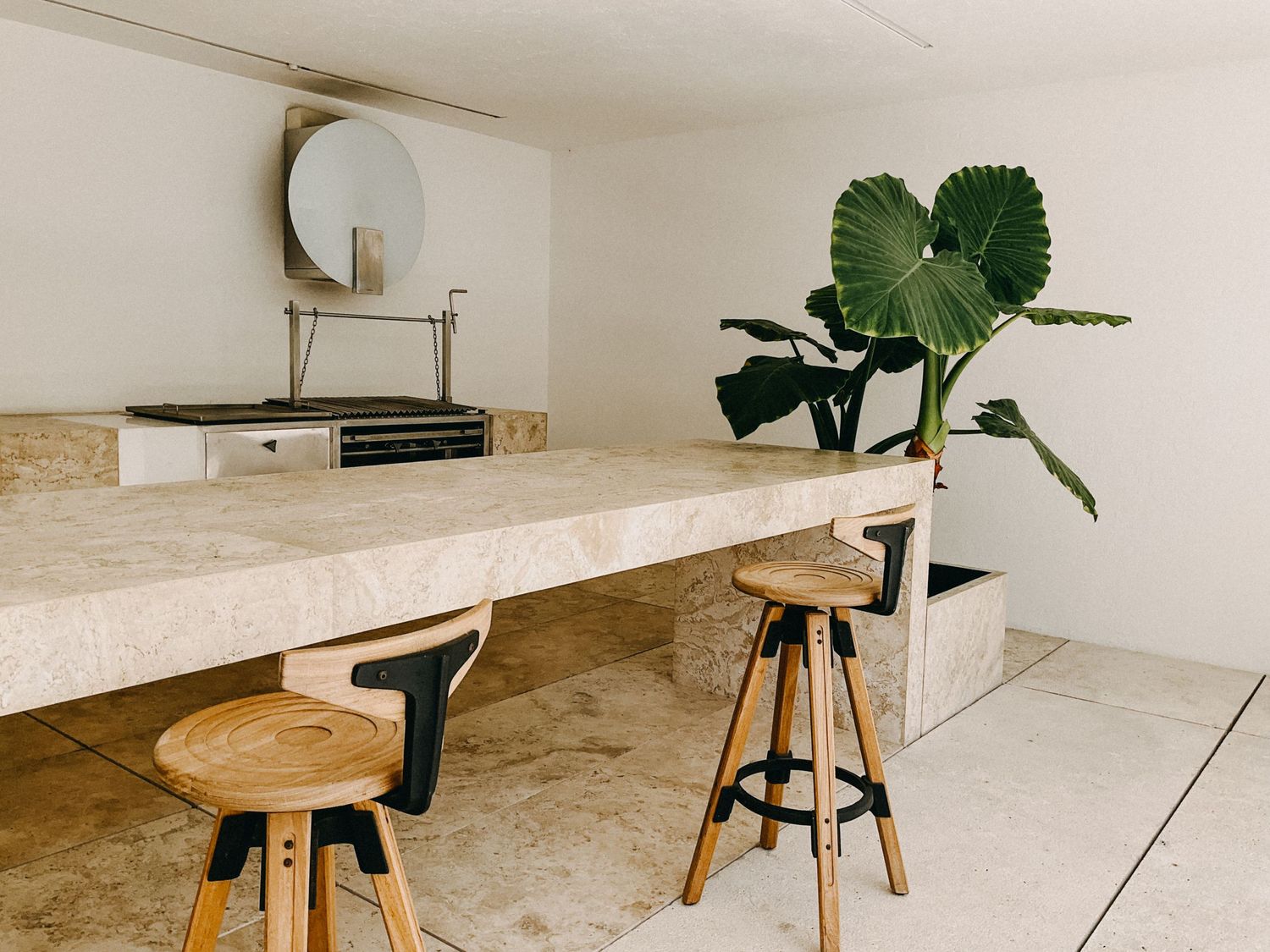Minimalist kitchen bar with travertine countertop, wooden stools, round mirror, and tropical plant.