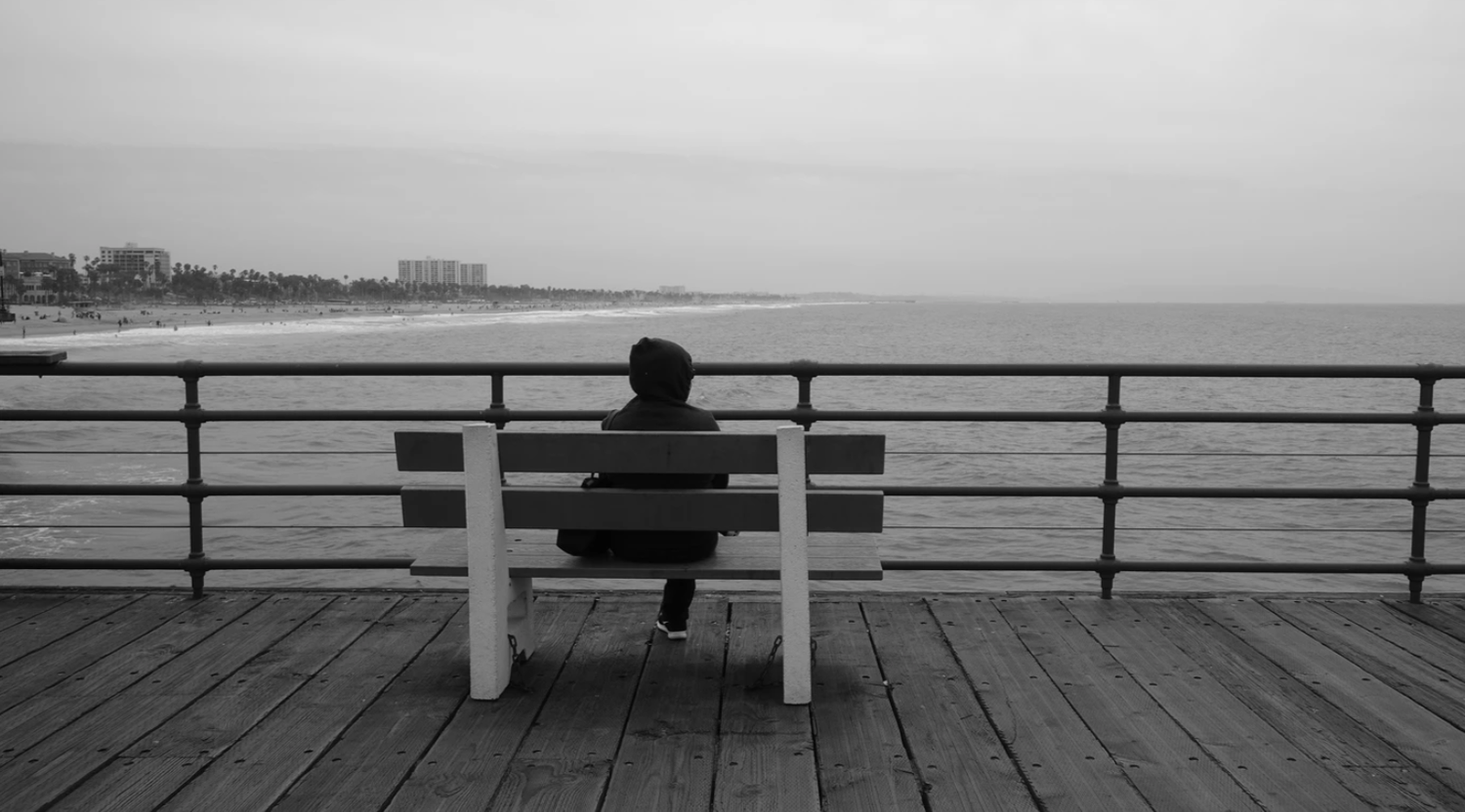 Person sitting alone on a bench on a wooden pier overlooking a calm ocean beach on an overcast day.