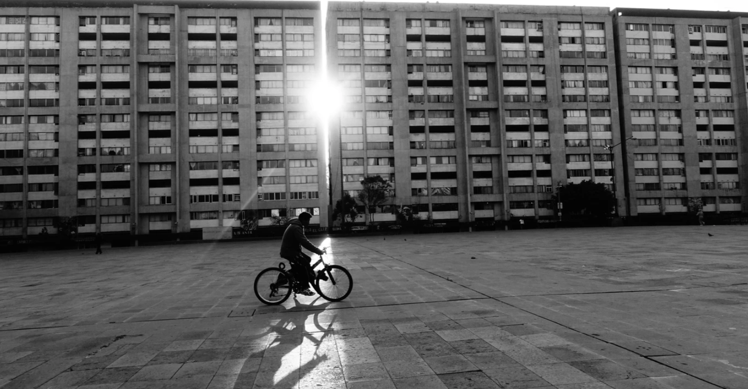 Black and white photo of a cyclist riding across a vast plaza in front of a large brutalist building with dramatic sunlight.