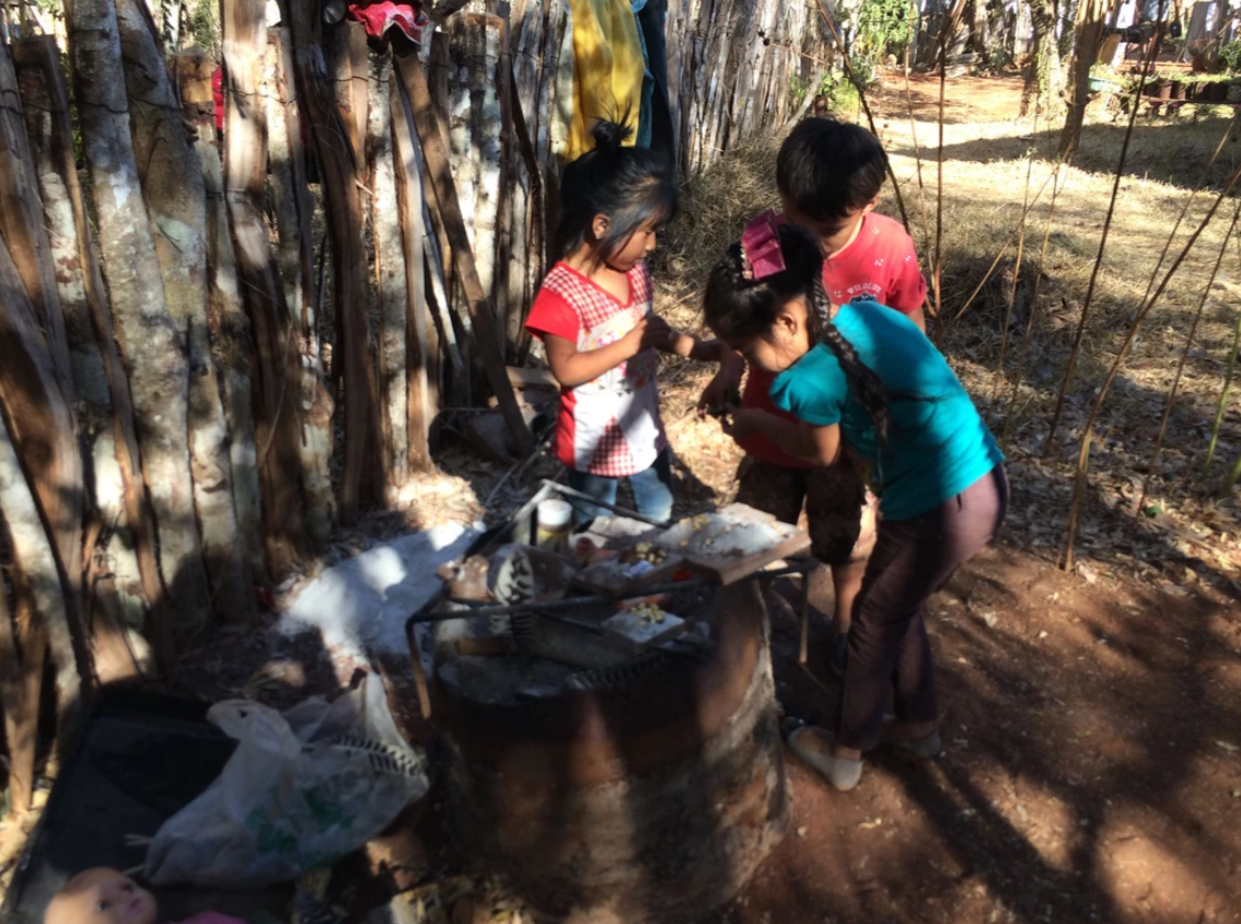 Children gathered around an outdoor fire pit in a wooded area in a rural setting.