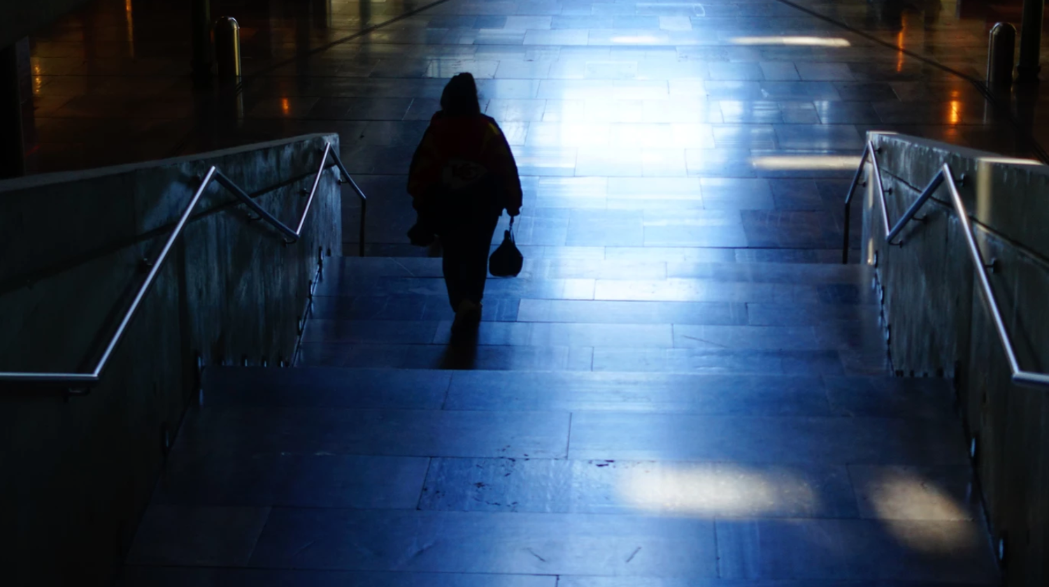 Silhouetted person walking up dimly lit blue-toned staircase with metal railings in an urban setting.