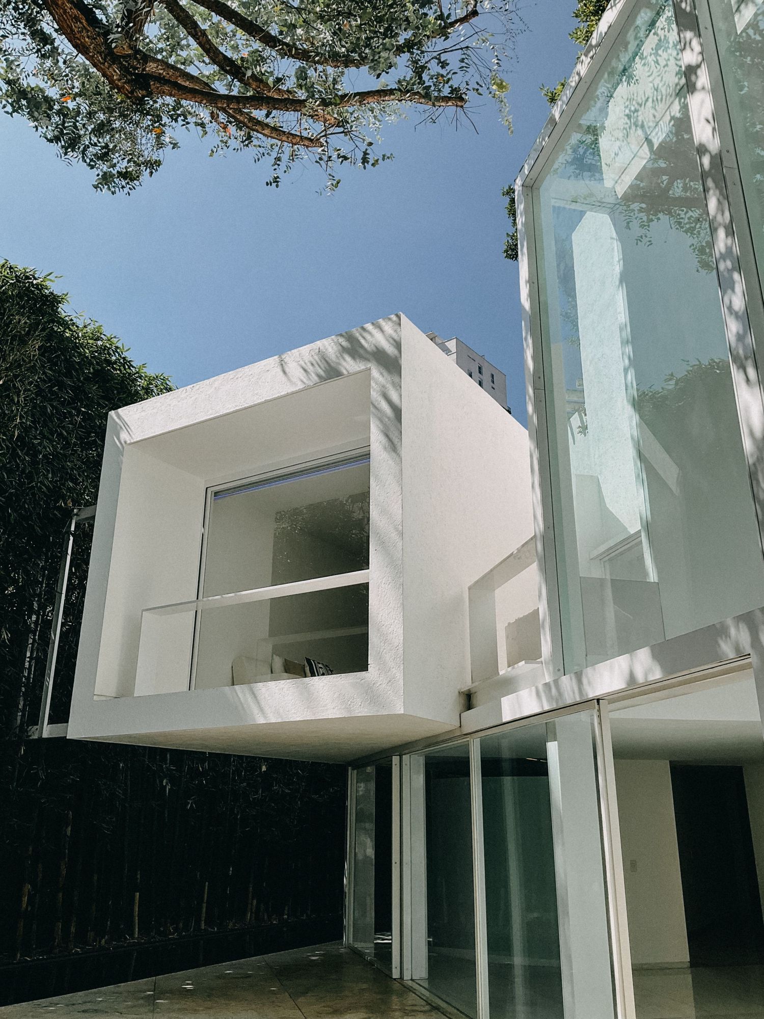 Modern white cubic architecture with large glass panels and cantilevered upper floor under blue sky.