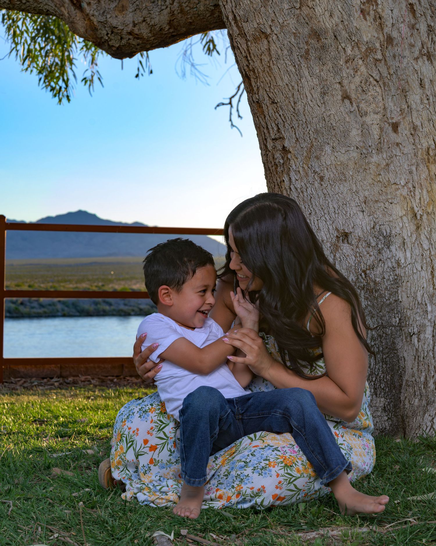 Mother and young son share a joyful moment laughing together under a tree near a scenic waterway.