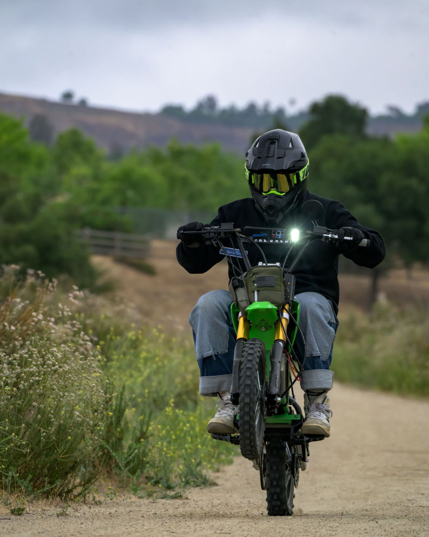 Rider in black helmet and goggles on a green dirt bike riding down a gravel trail surrounded by greenery.
