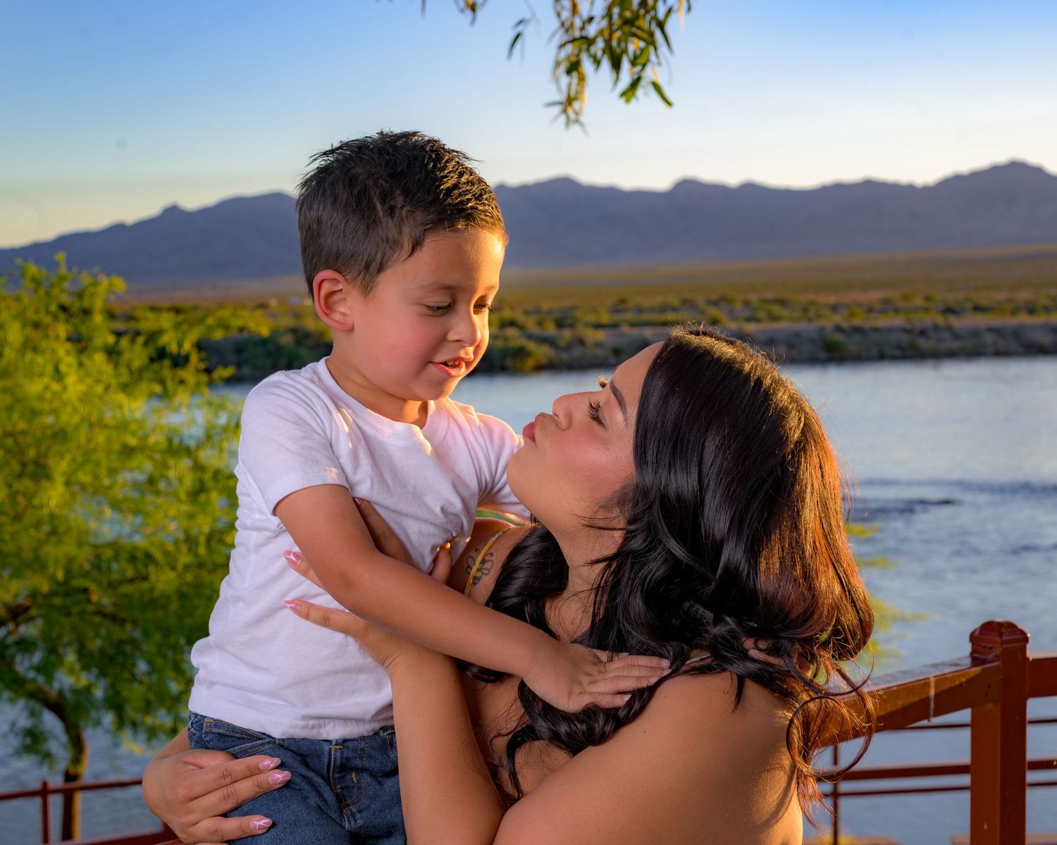 A mother holds her young son outdoors near a river with mountains at sunset, both smiling joyfully.