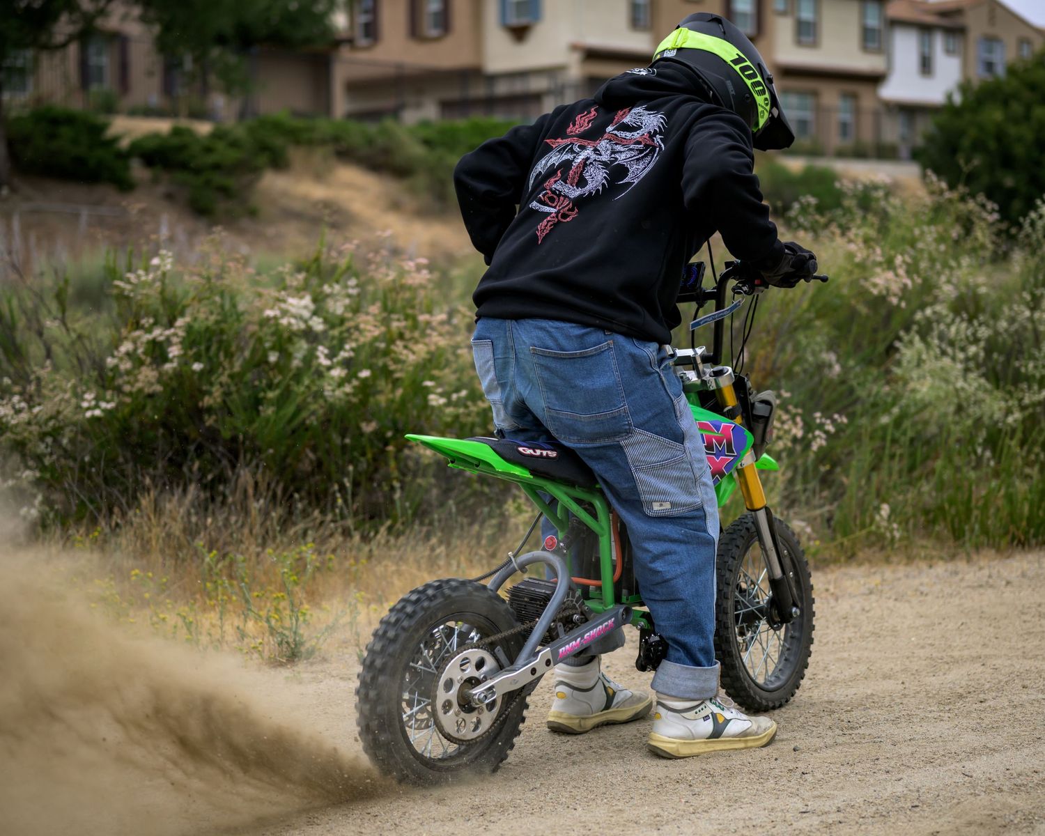 A rider in a black hoodie and jeans performs a wheelie on a green dirt bike on a dusty trail.