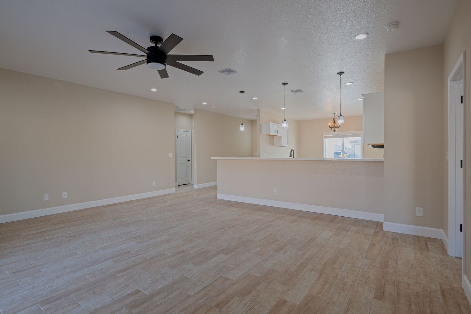 Empty room with beige walls, hardwood flooring, ceiling fan, and pendant lights in an open concept layout.