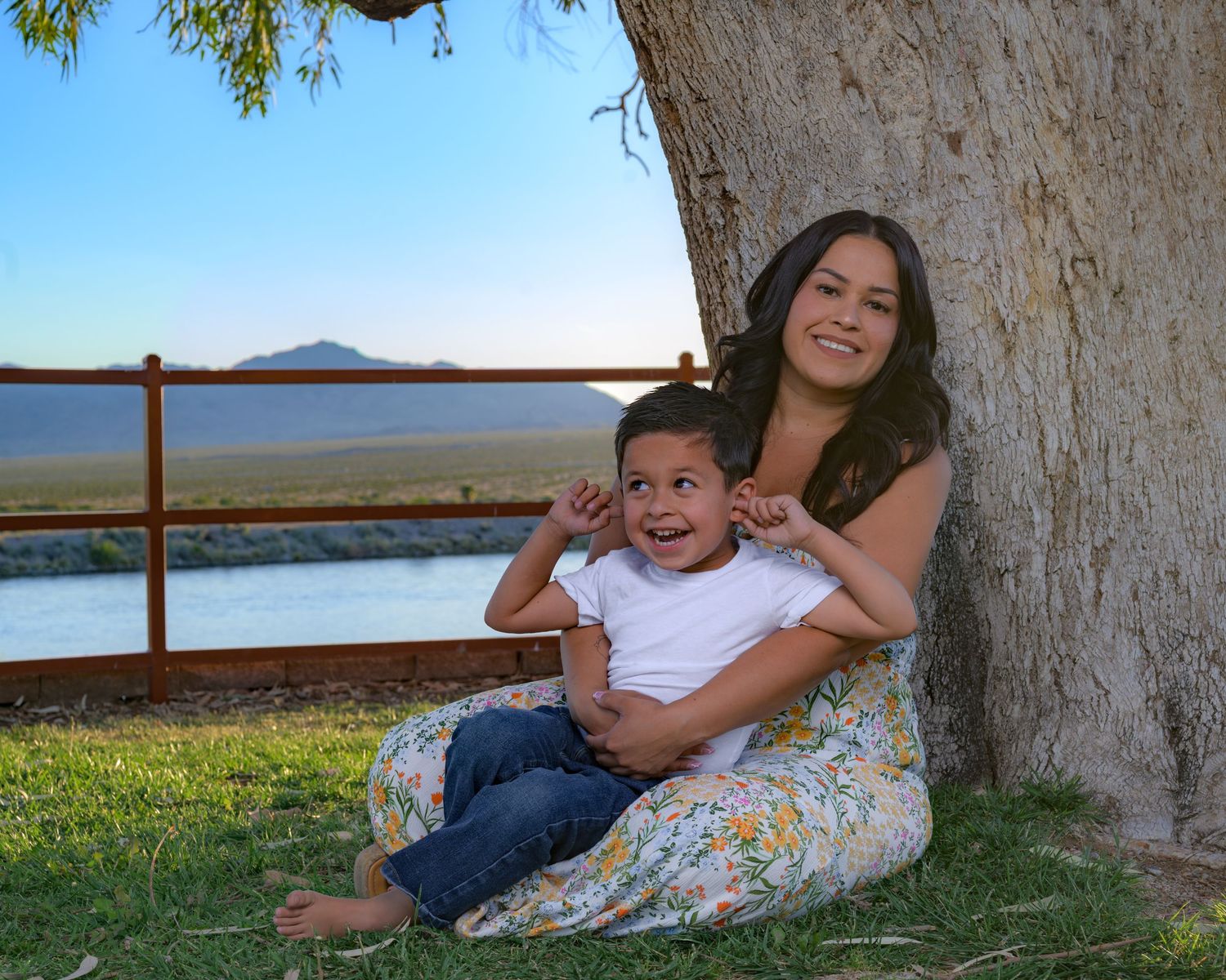 A smiling woman and young boy sit together under a large tree with scenic mountain views behind them.