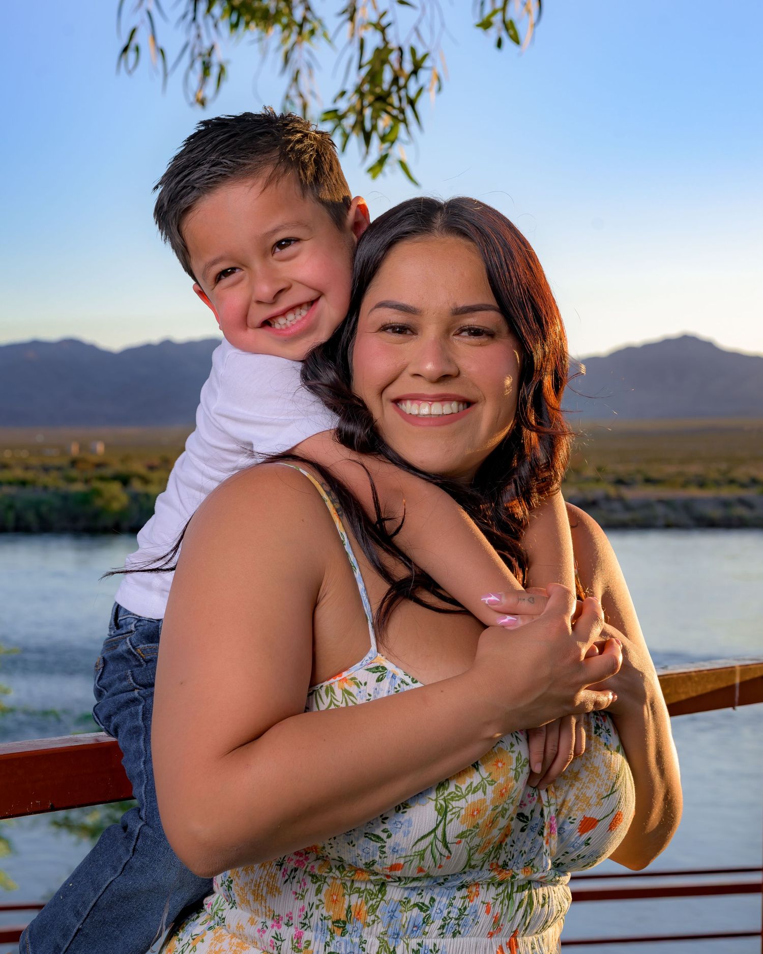 A smiling woman in a floral dress holds a happy young boy on her back near a scenic mountain lake.