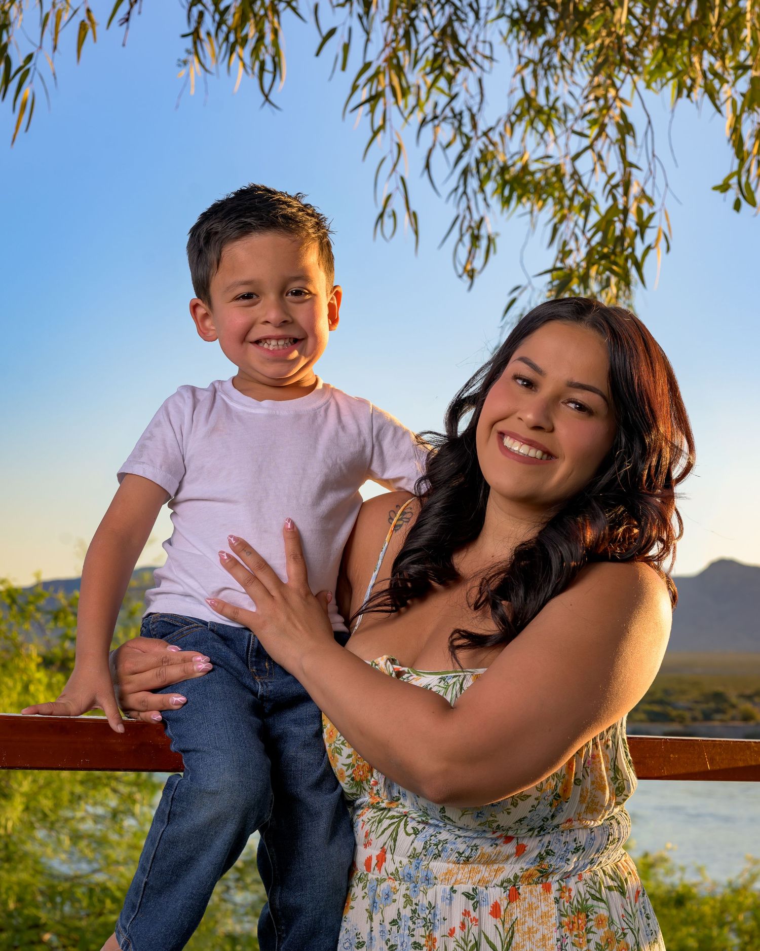 A cheerful boy sits on a railing held by a smiling woman in a floral dress with a scenic outdoor backdrop.