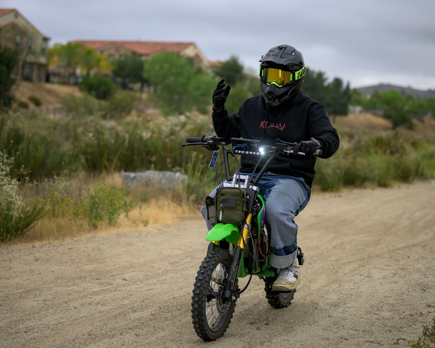 Rider in green helmet waves while riding a green dirt bike on a dusty trail with houses in background.