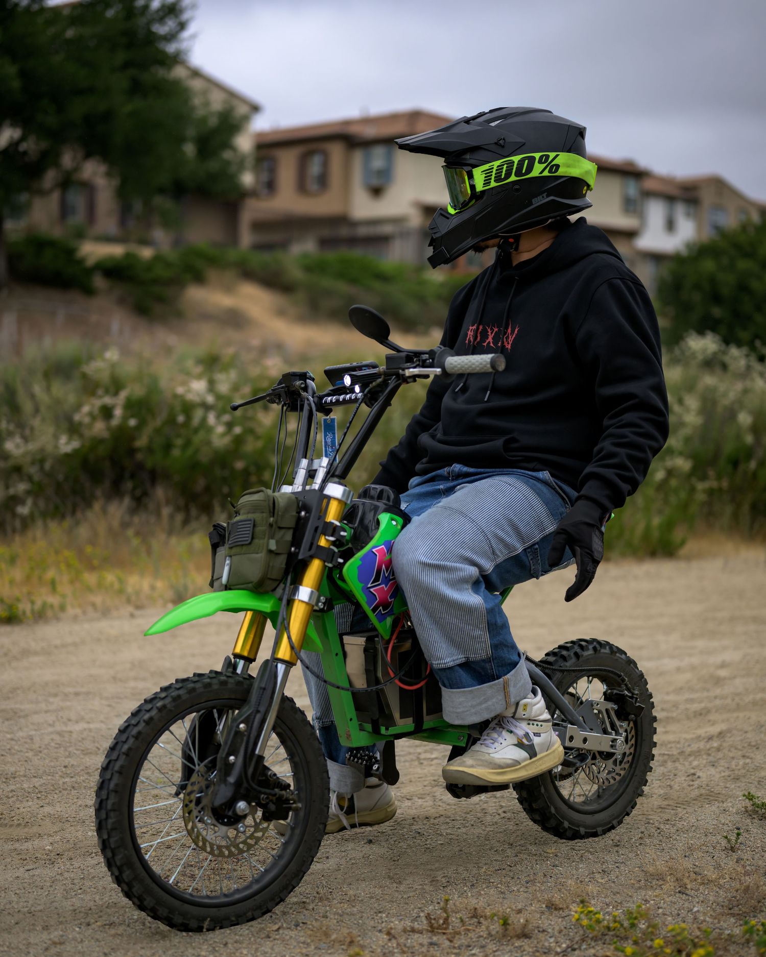Young rider in black hoodie and green helmet sits on a green dirt bike on a dirt trail near suburban homes.