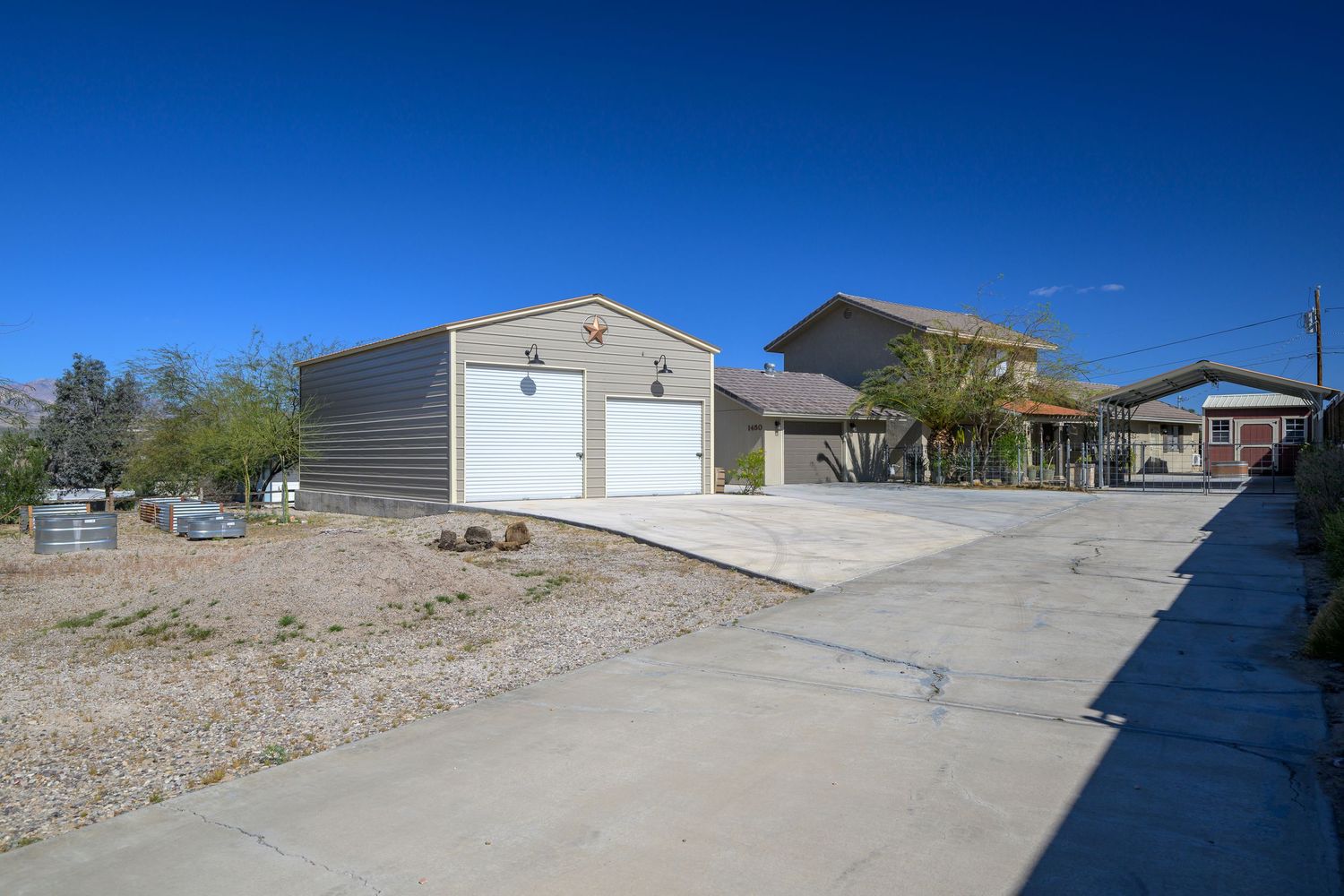Gray metal garage building with concrete driveway and covered carport structure in a rural desert setting.