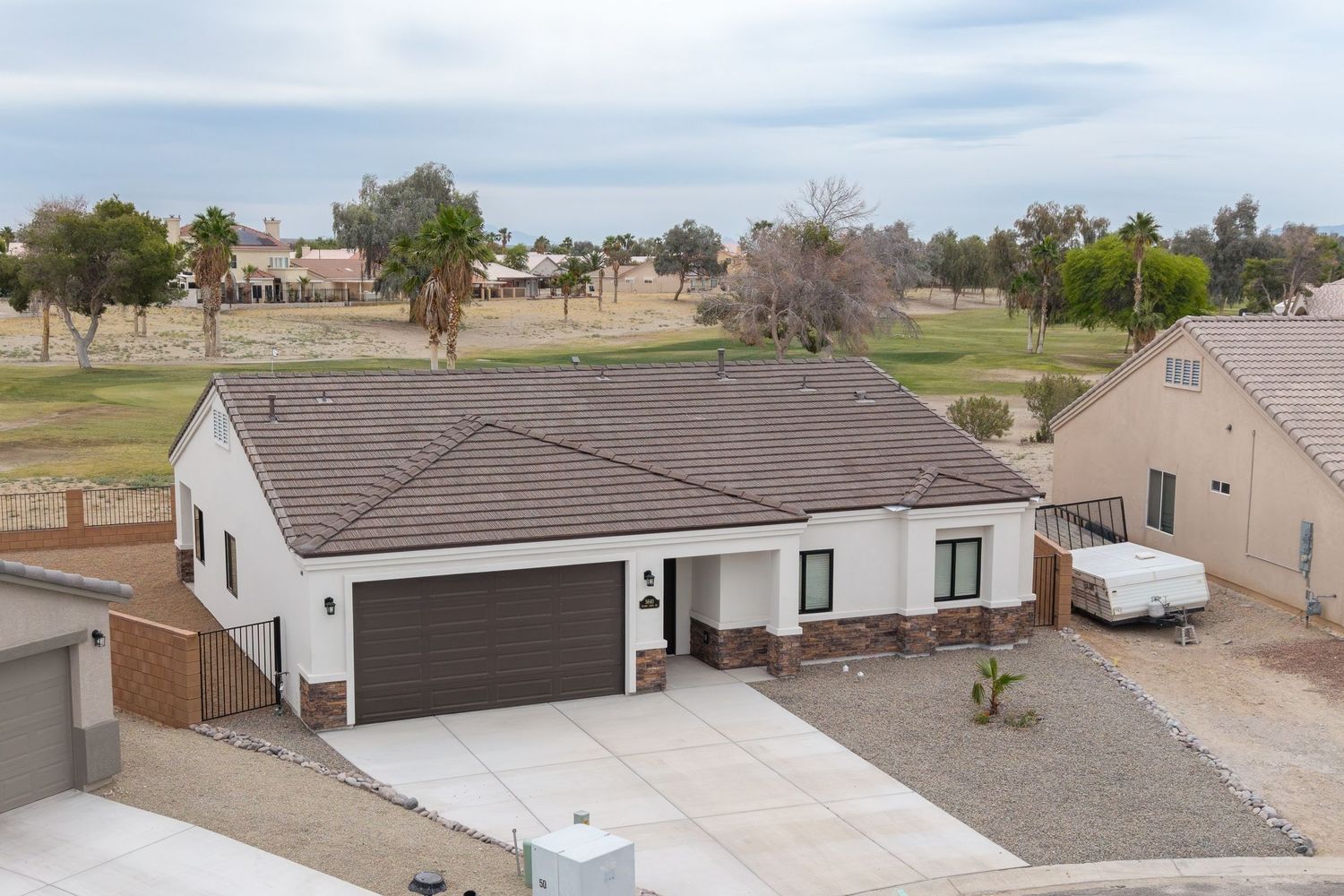 Aerial view of a white single-story home with brown tile roof, two-car garage, and desert landscaping near a golf course.