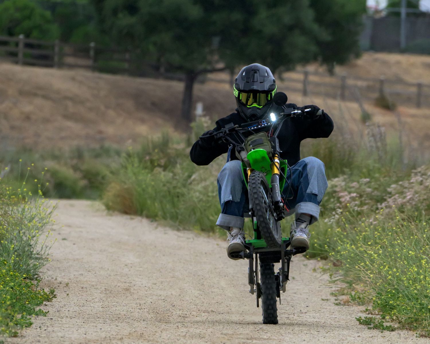 Motorcyclist performing a wheelie on a green dirt bike along a rural gravel path wearing a helmet and protective gear.