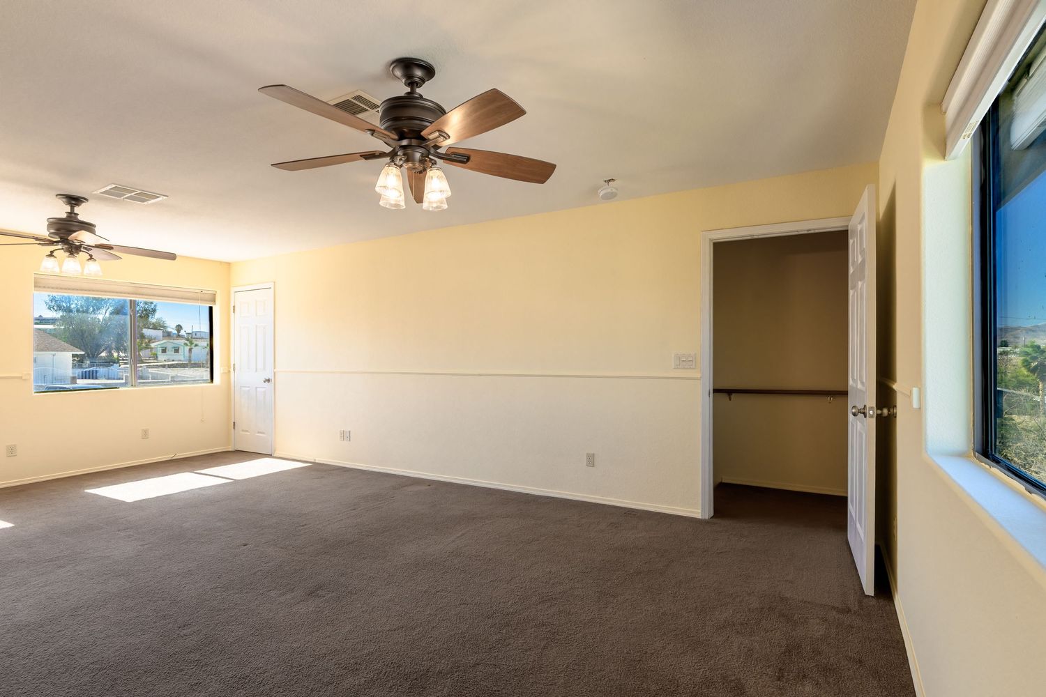 Empty beige room with brown carpet flooring, ceiling fans, and natural light coming through windows.