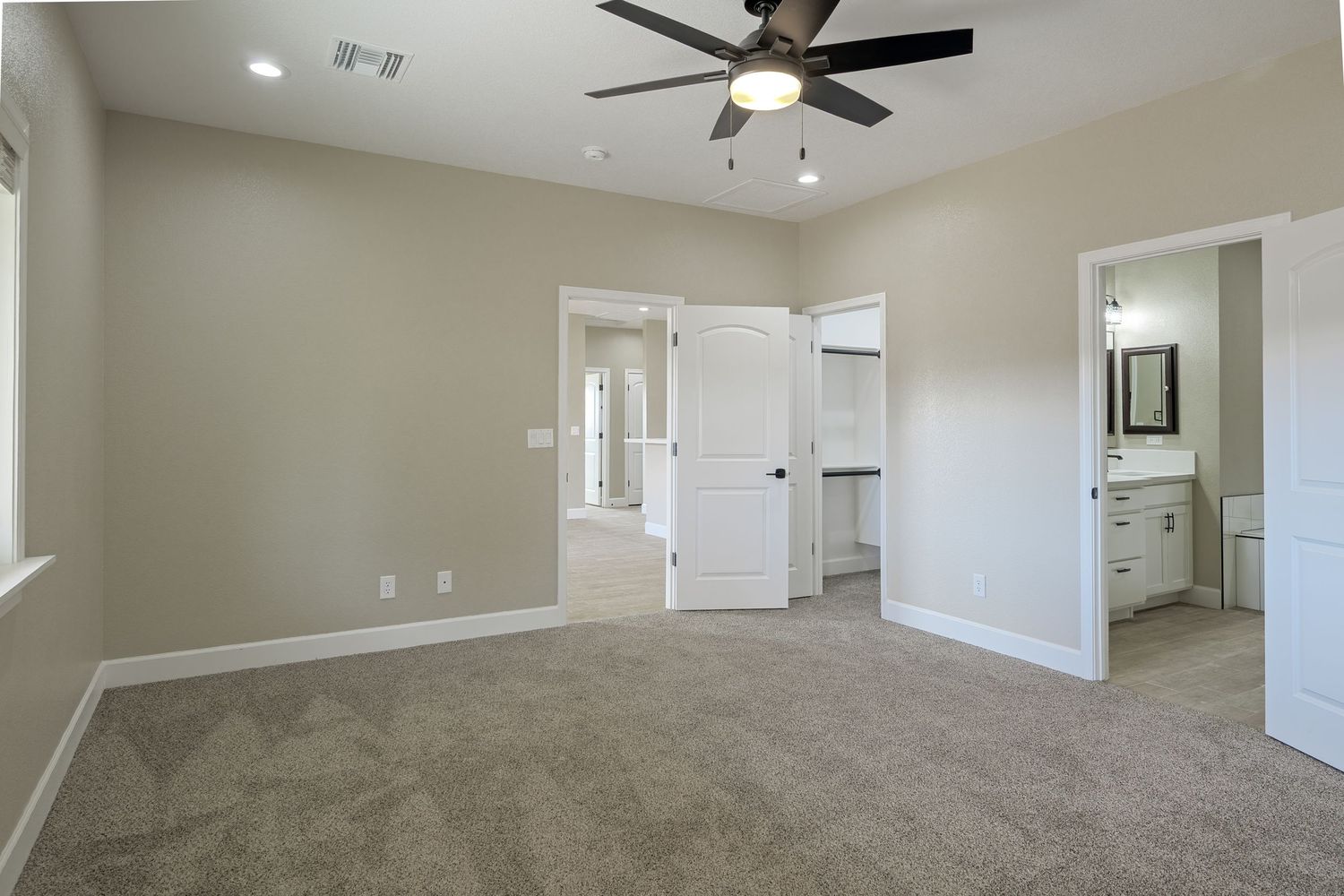 Empty bedroom with beige carpet, tan walls, ceiling fan, and white doors leading to bathroom and closet areas.