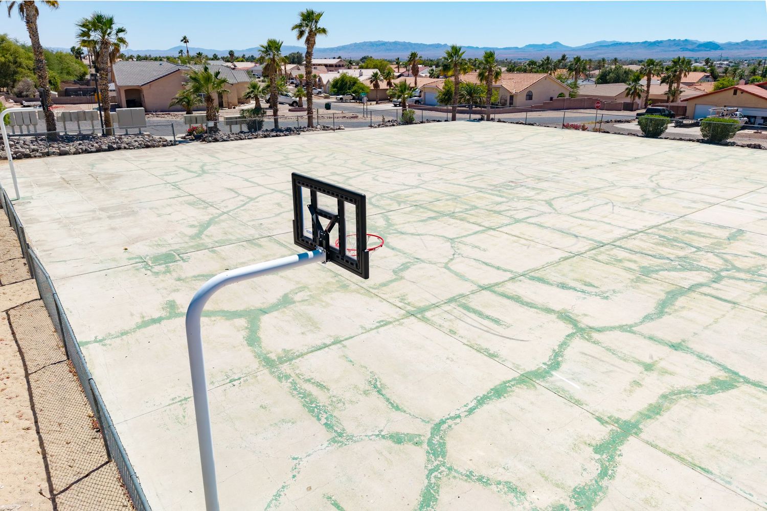 Outdoor basketball court with a single hoop, cracked concrete surface, palm trees, and mountain views in the background.