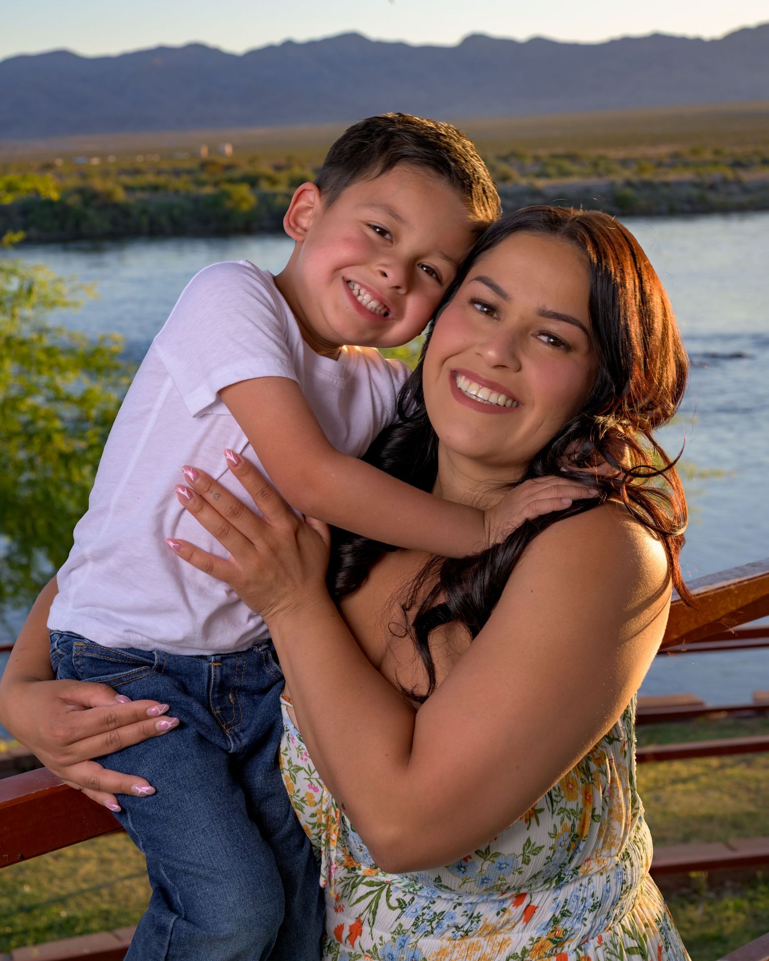 A young boy hugs a smiling woman in a floral dress outdoors by a river with mountains in the background.