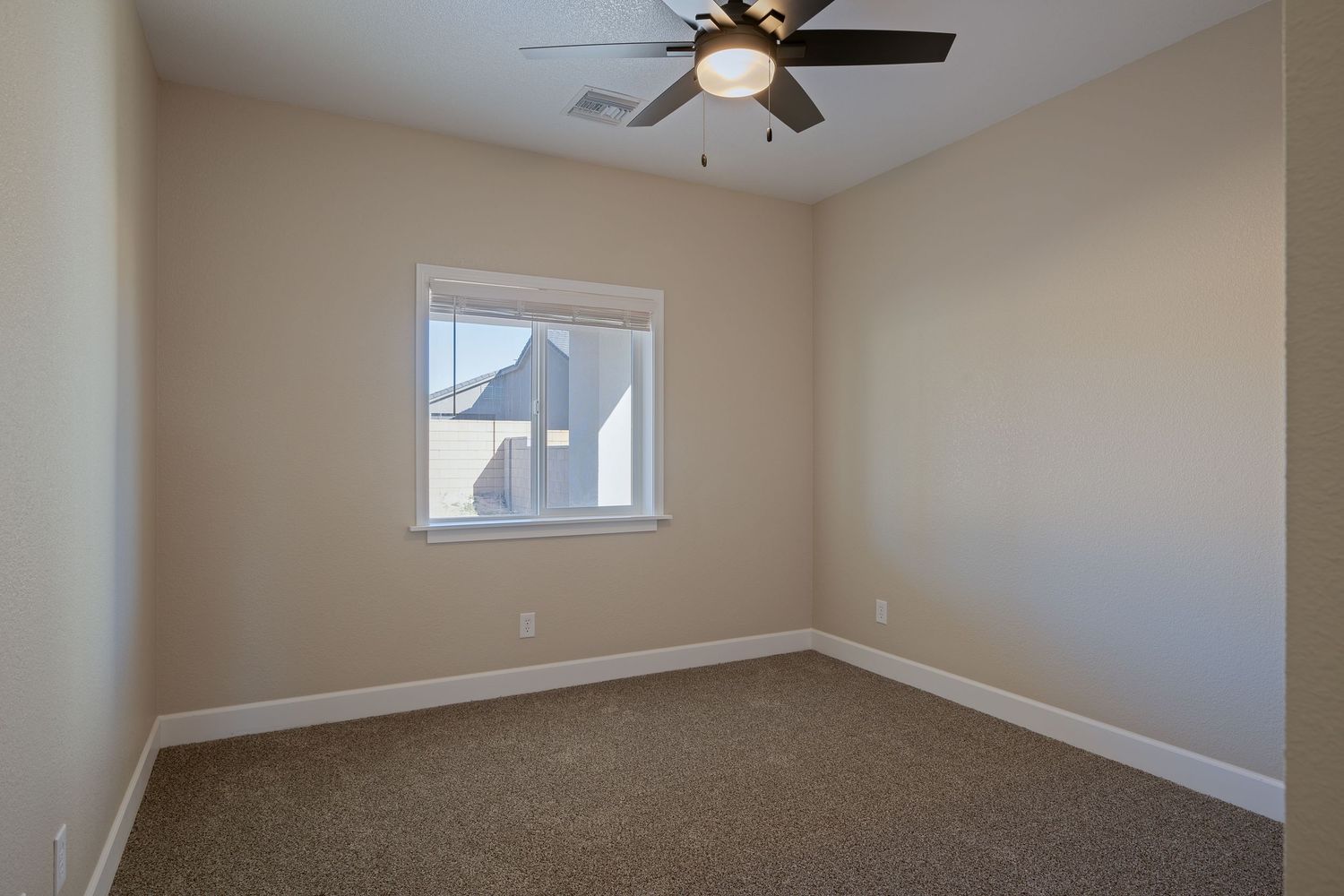 Modern bedroom with beige walls, carpeted floor, ceiling fan and single window casting geometric shadows.