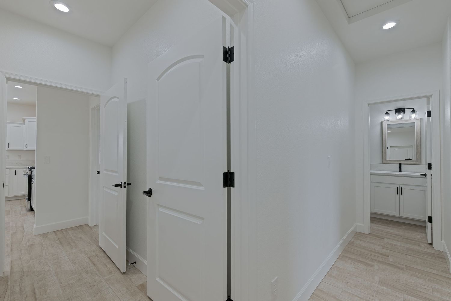 Bright hallway with white double doors, light wood floors, and a glimpse of a modern bathroom vanity with mirror.