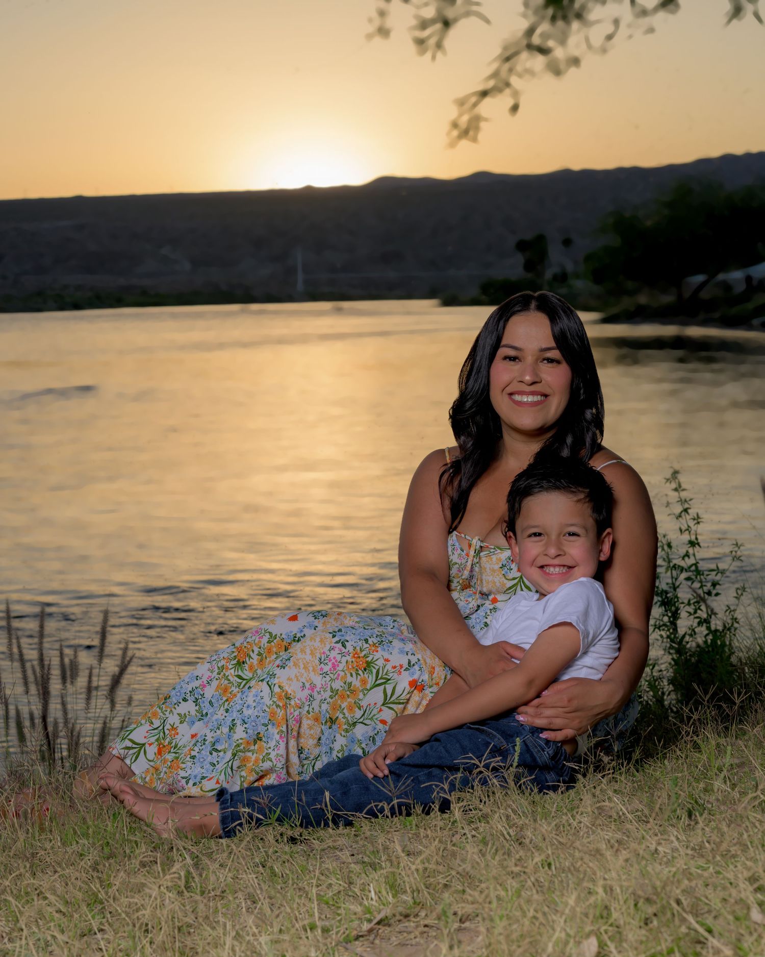 Mother and son smiling by a lake at golden sunset in a floral dress.
