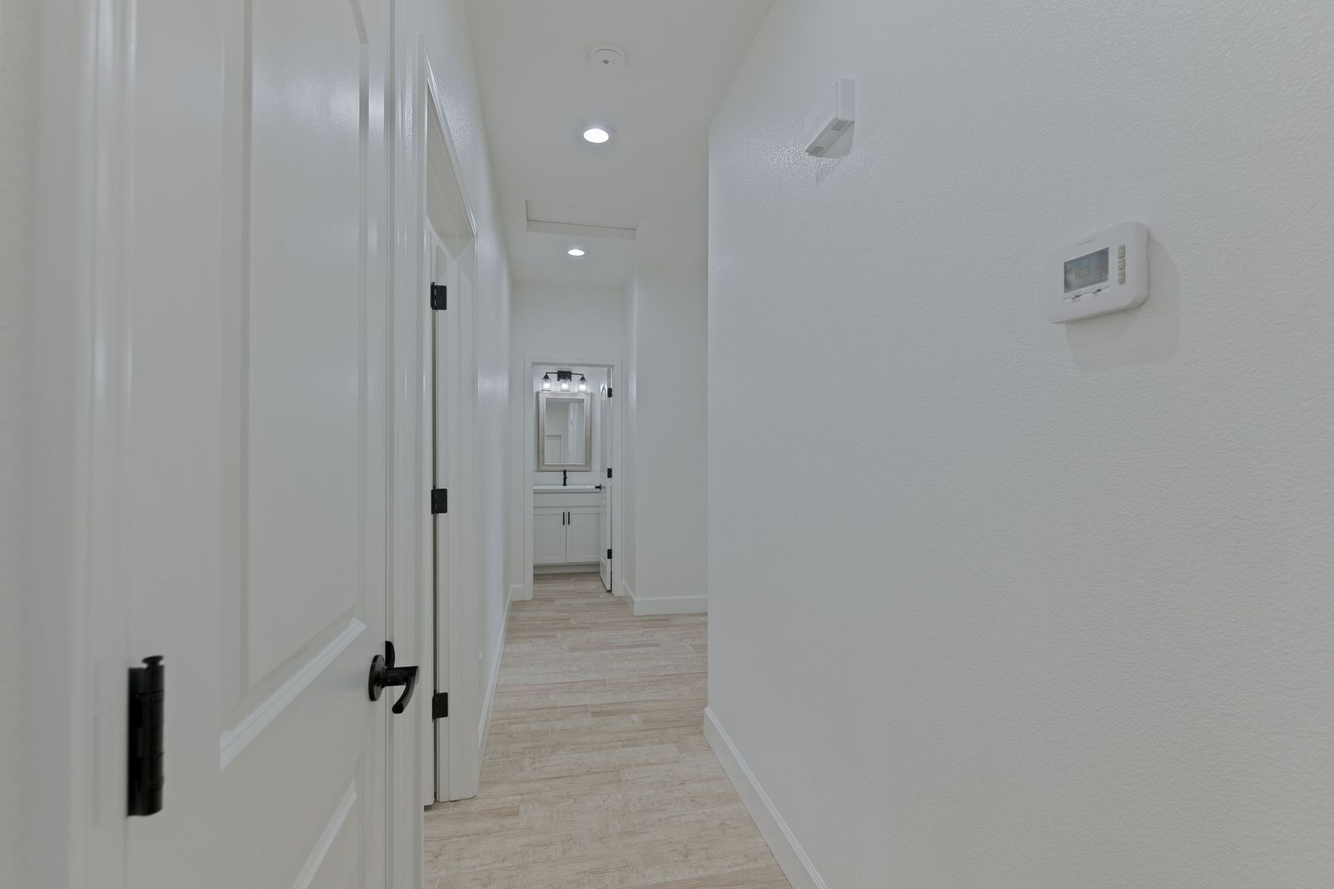 Bright white hallway with light wood floors, black door hardware, recessed lighting, and a thermostat on the wall.