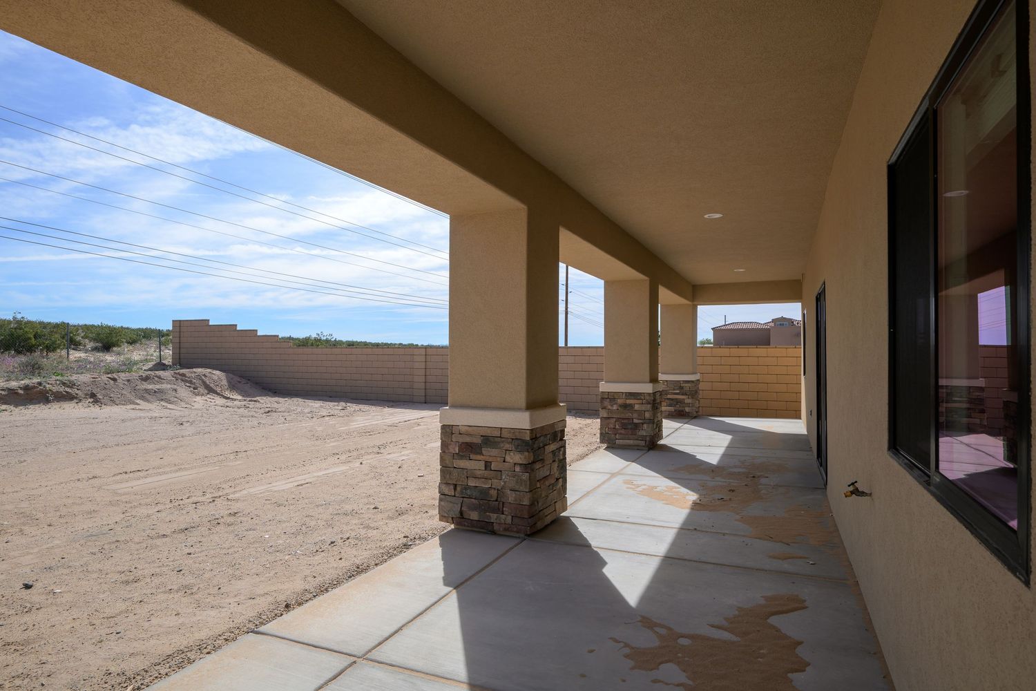 Covered patio with stone accents and concrete walkway extends along desert home exterior under blue sky.