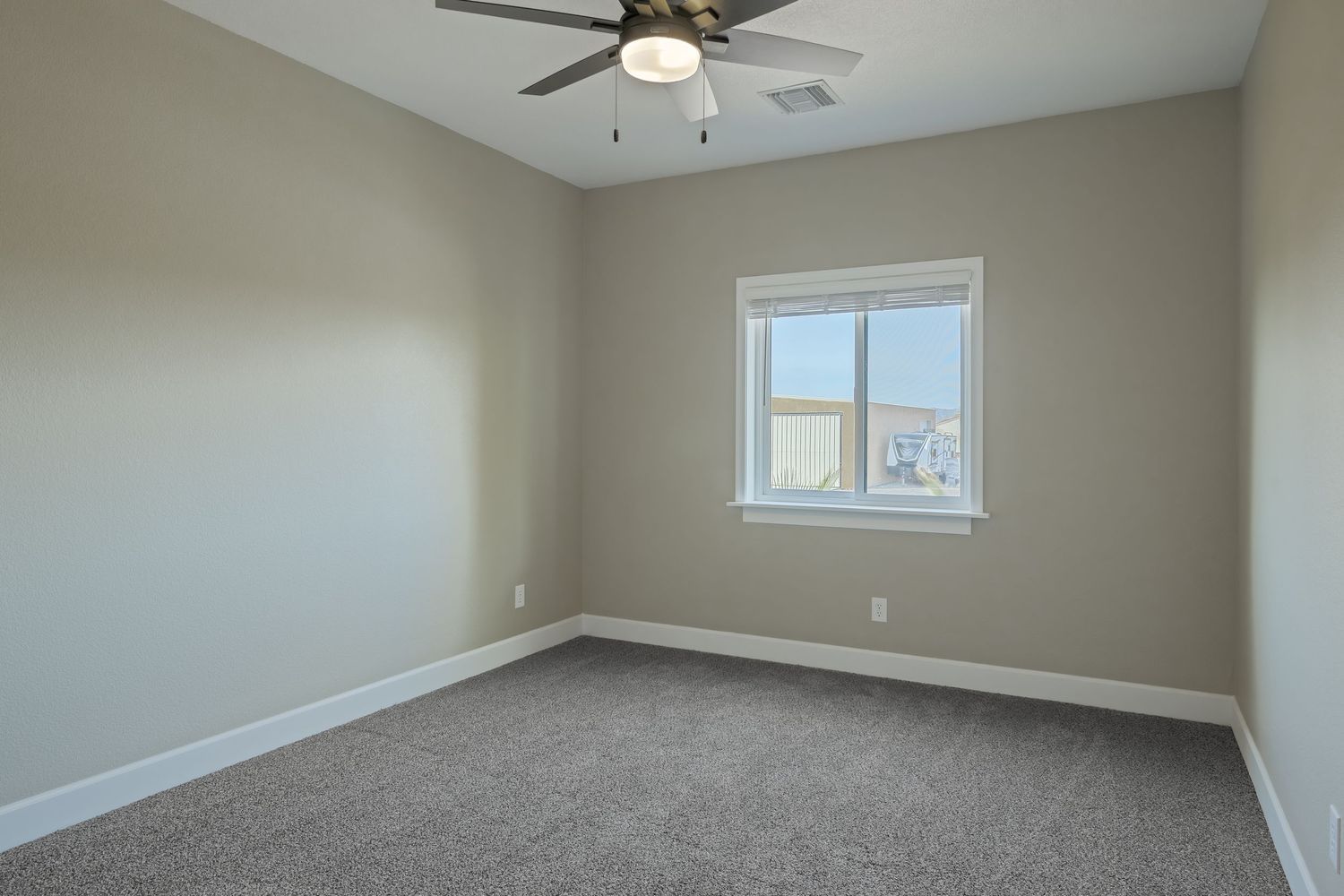 Empty room interior with beige walls, gray carpet flooring, and a ceiling fan with windows providing natural light.