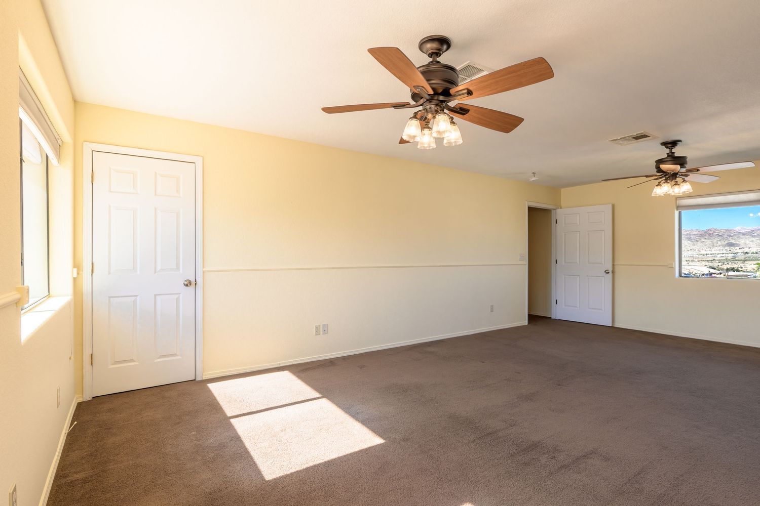 Spacious empty room with beige walls, ceiling fans, and natural sunlight streaming through windows.