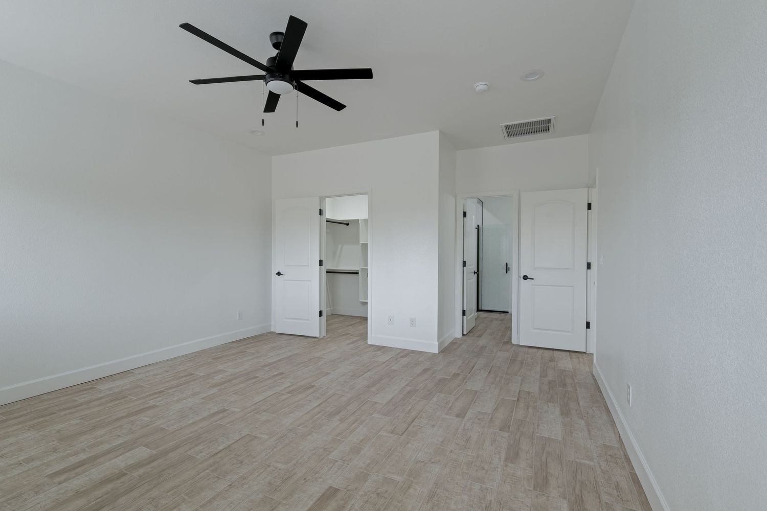 Bright empty bedroom with light wood floors, white walls, black ceiling fan, walk-in closet, and two doors.