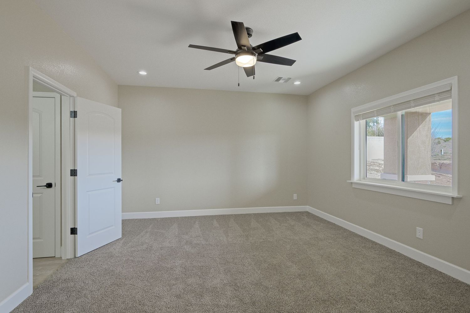 Empty bedroom with beige carpet, white trim, ceiling fan and window showing exterior brick wall and blue sky.
