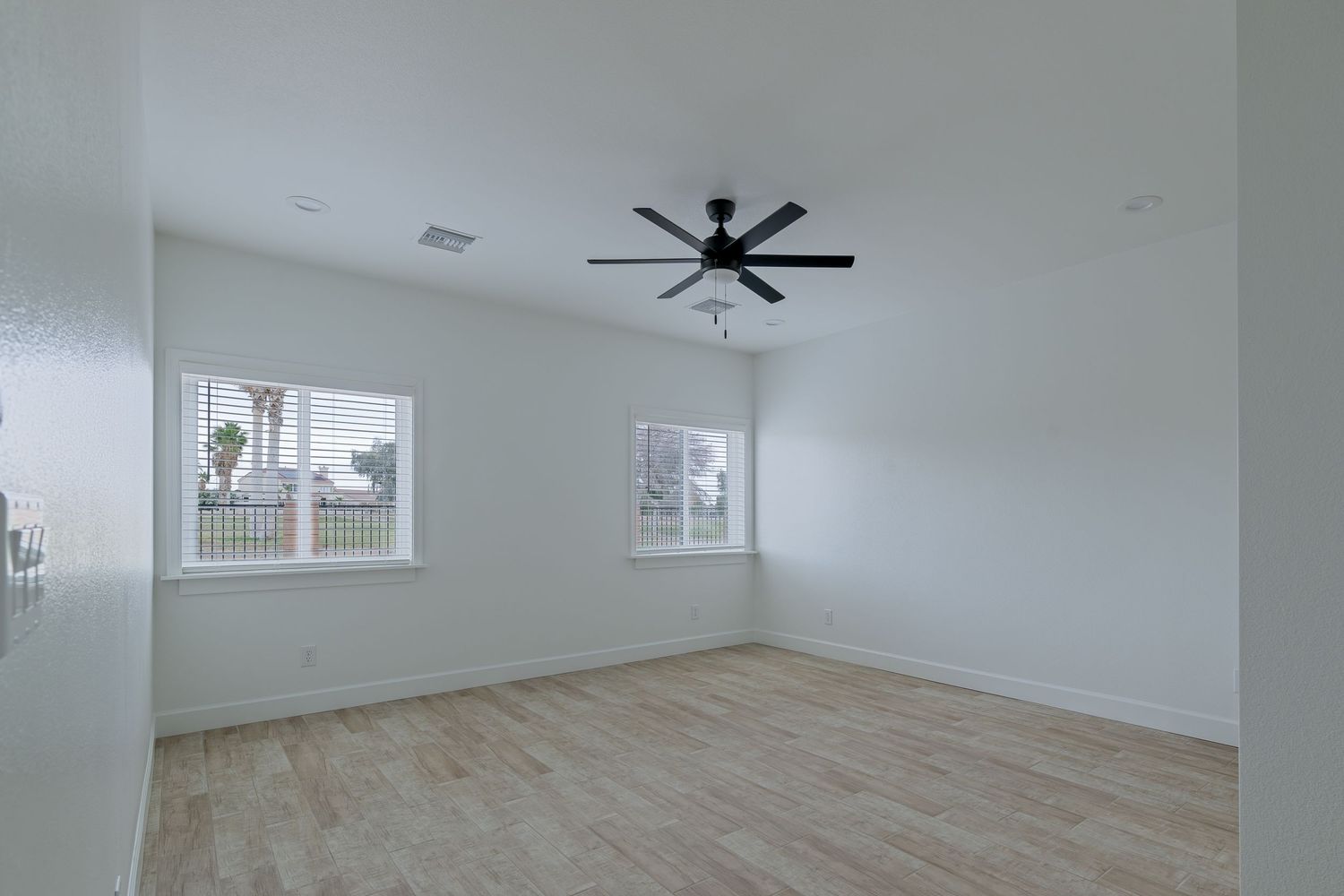 Bright empty room with light wood floors, white walls, and a black ceiling fan above a window with natural light.