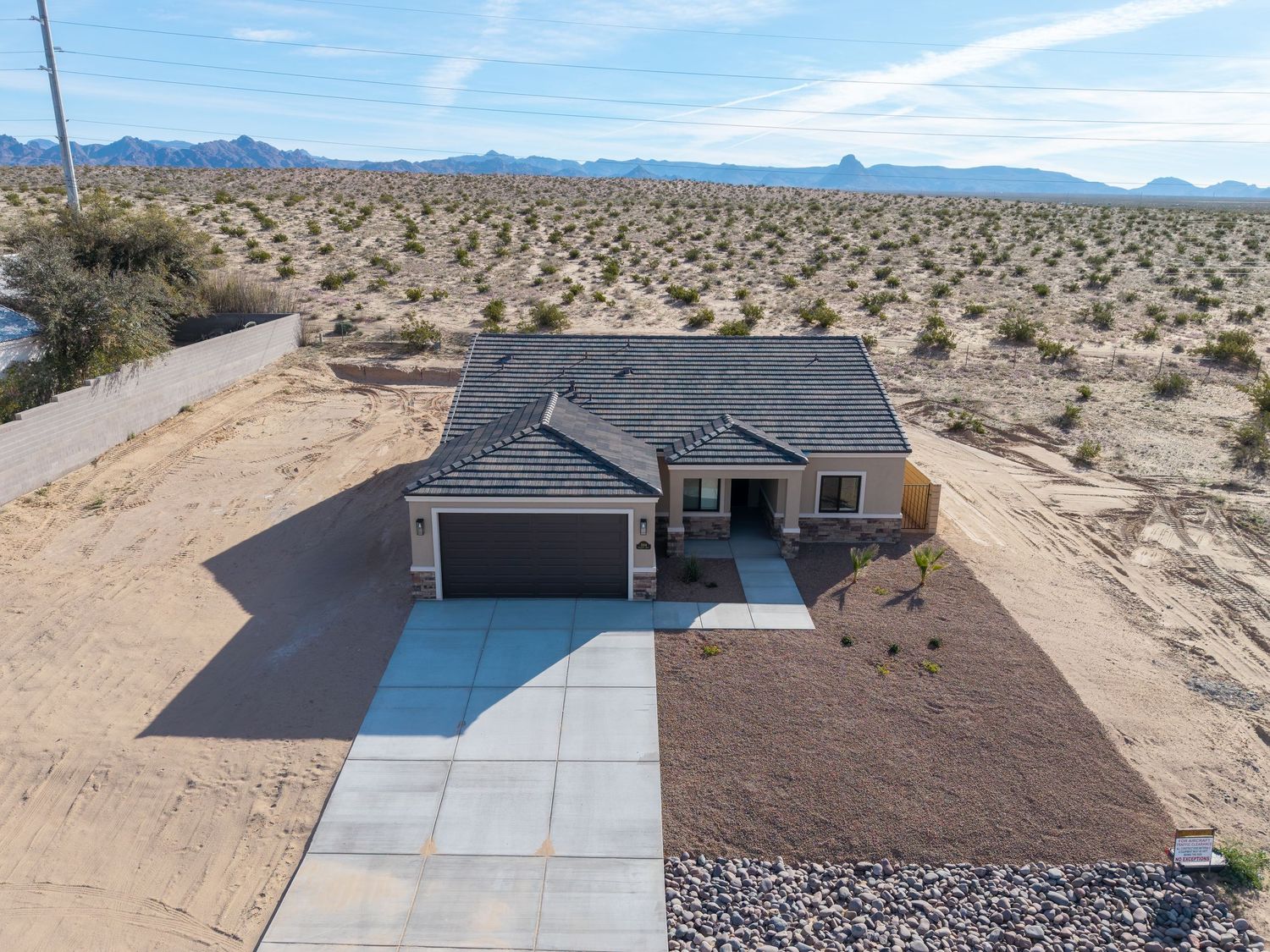 Aerial view of modern single-story house with gray roof and attached garage in desert landscape with mountains in background.
