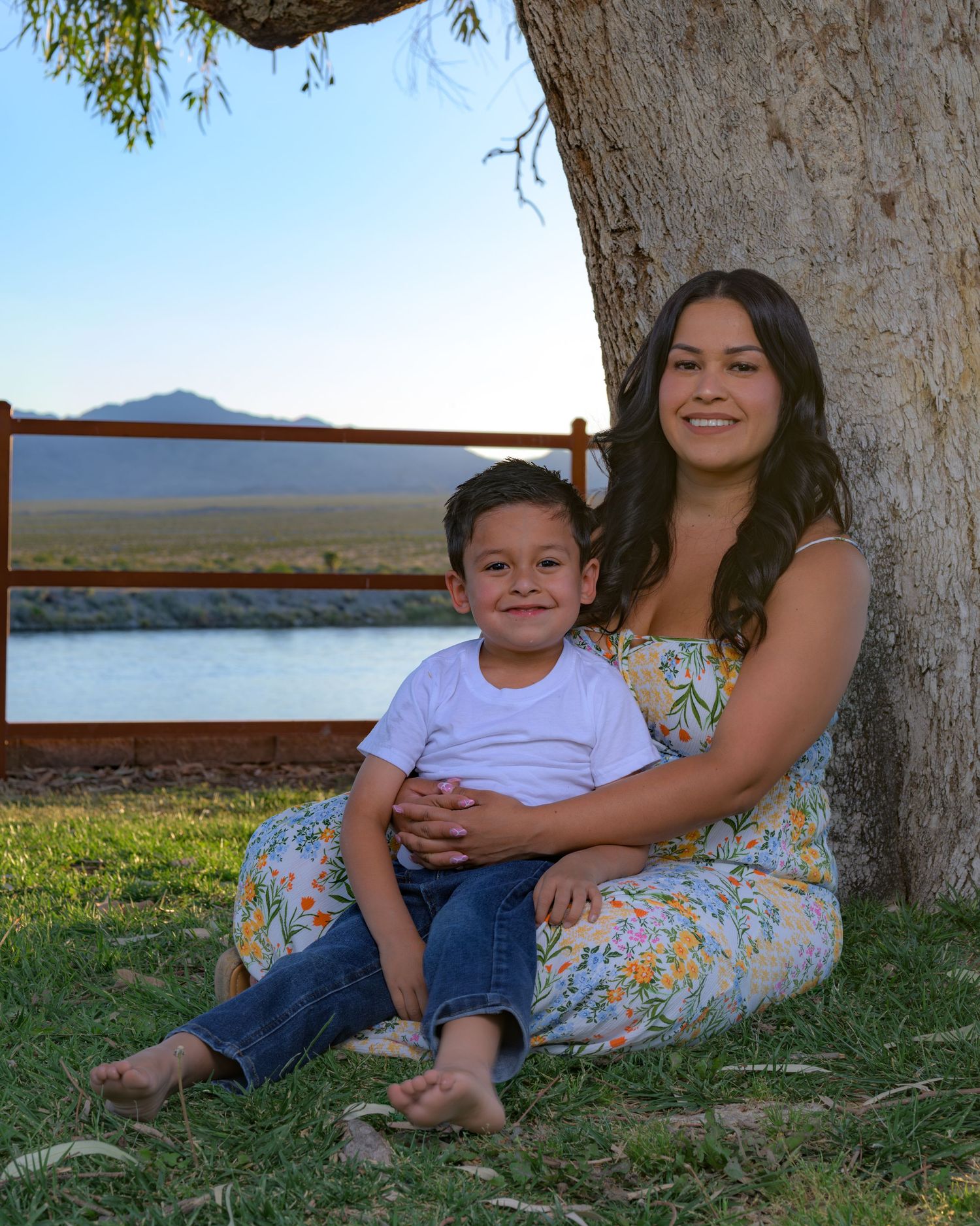 A young woman in a floral dress sits with a smiling boy under a tree by a scenic lake and mountains.