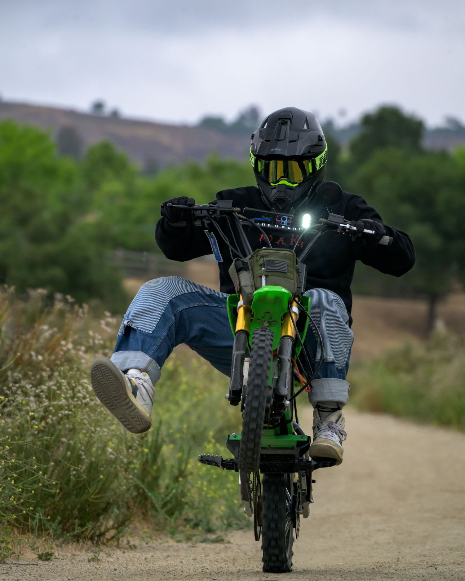 Rider in green helmet does a wheelie on a green dirt bike on a rural path surrounded by lush greenery.
