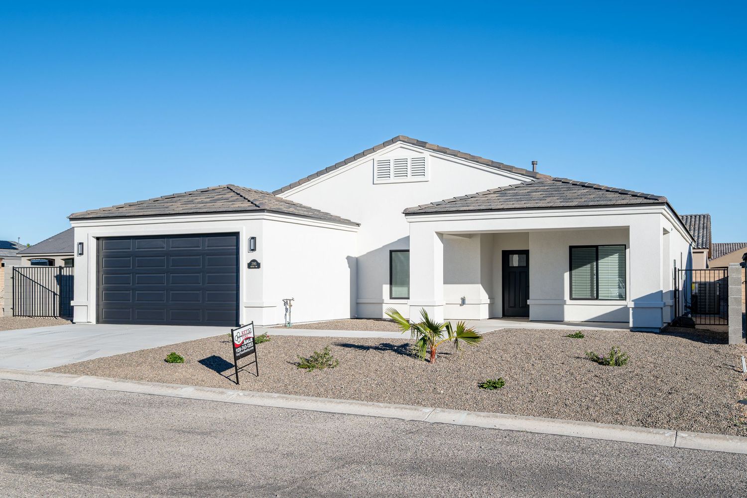 Modern residential homes with garages and desert landscaping line a suburban street under bright blue skies.
