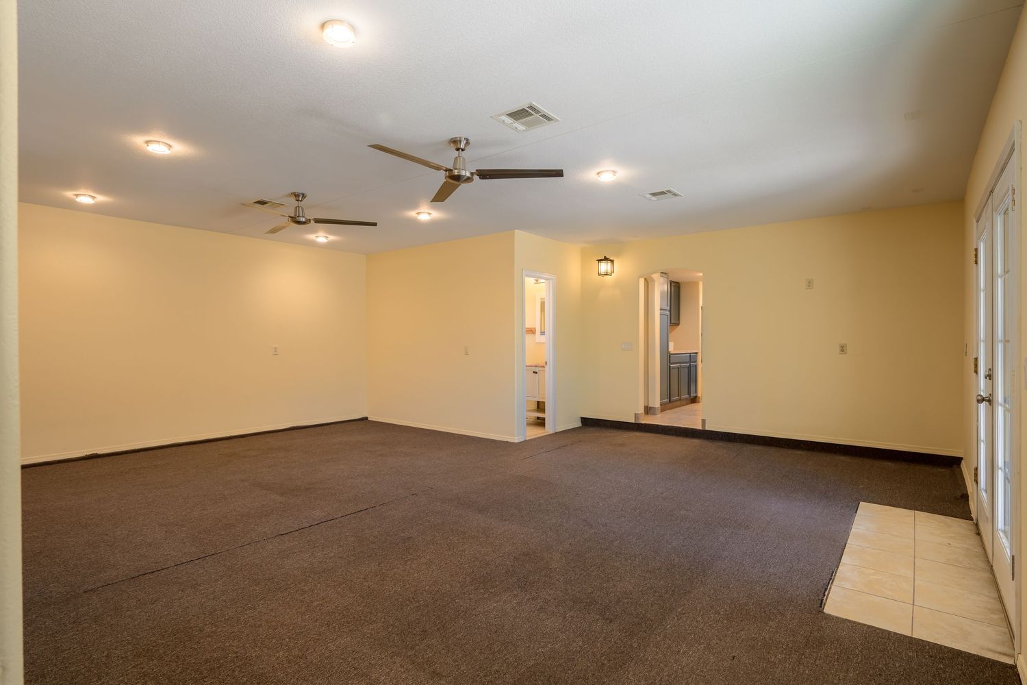 Large empty room with brown carpet flooring, beige walls, recessed lighting and ceiling fans in a commercial space.