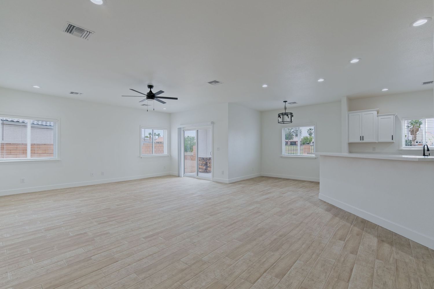 Bright open living room with light wood floors, ceiling fan, recessed lighting, and white walls leading to kitchen area.
