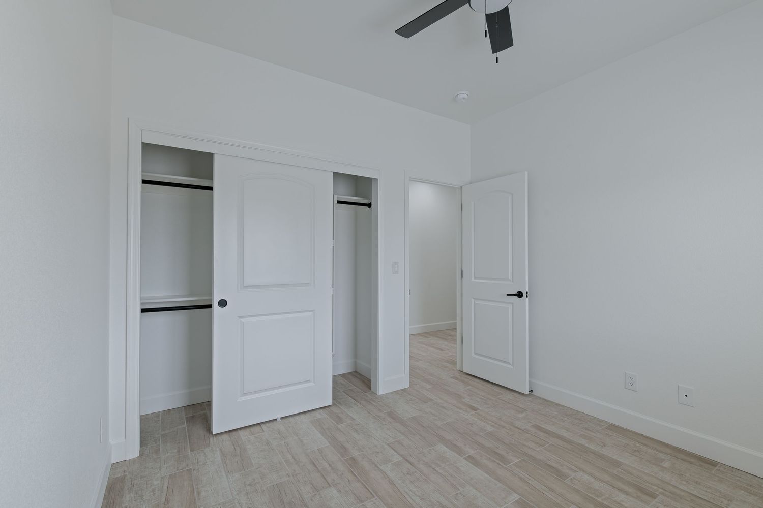 Bright bedroom with light wood floors, white walls, open closet with rod, and a ceiling fan in a modern home.