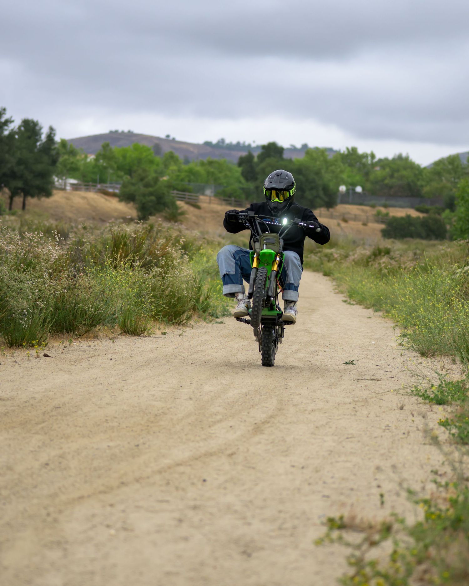 Rider on green dirt bike doing a wheelie on a dusty trail surrounded by green hills under cloudy skies.
