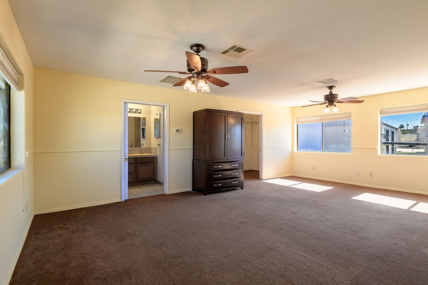 Empty beige room with brown carpet, ceiling fan, and windows showing multiple views of an unfurnished living space.