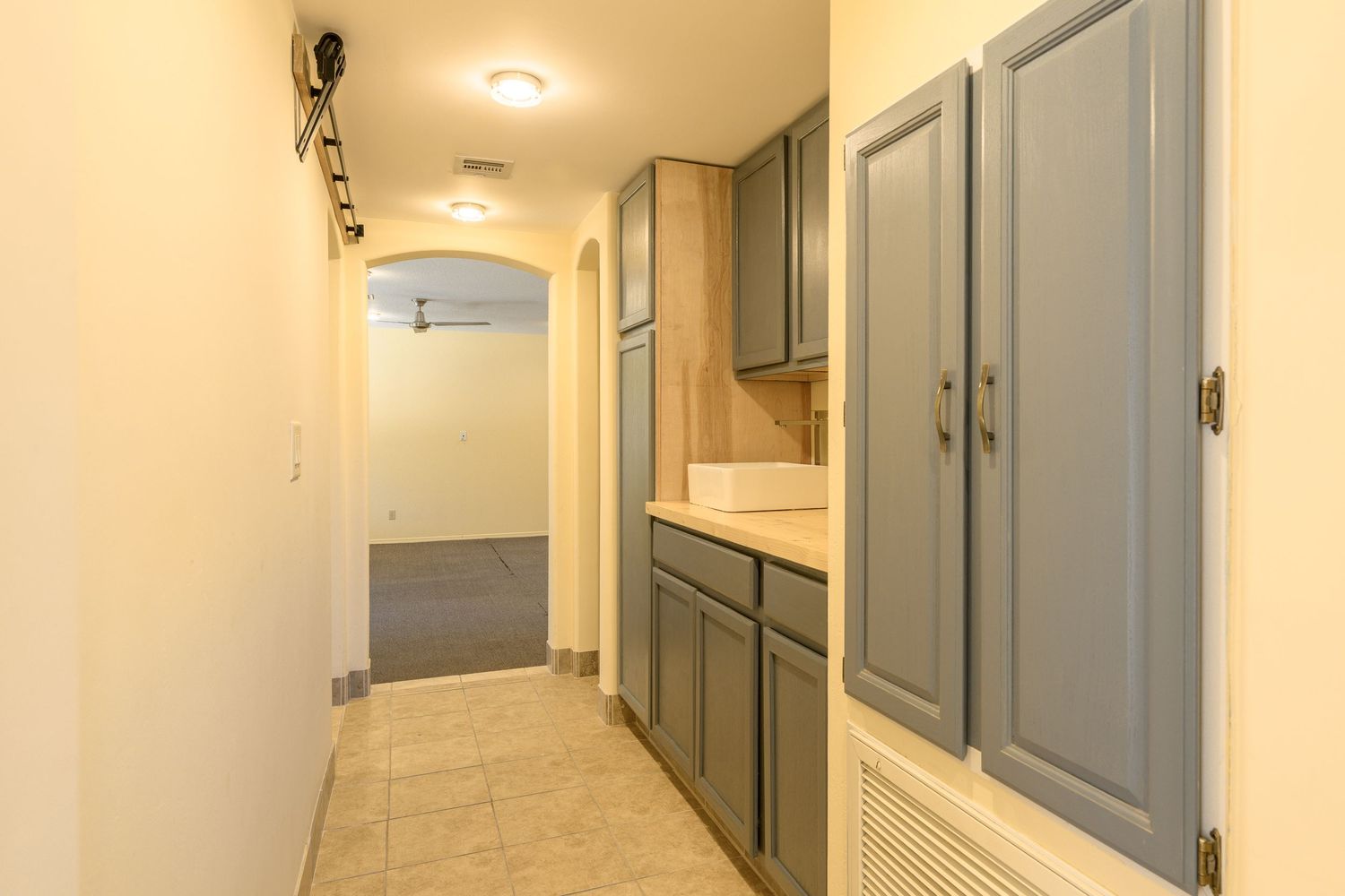 Long hallway with gray cabinets, tile flooring and cream walls leading to another room.