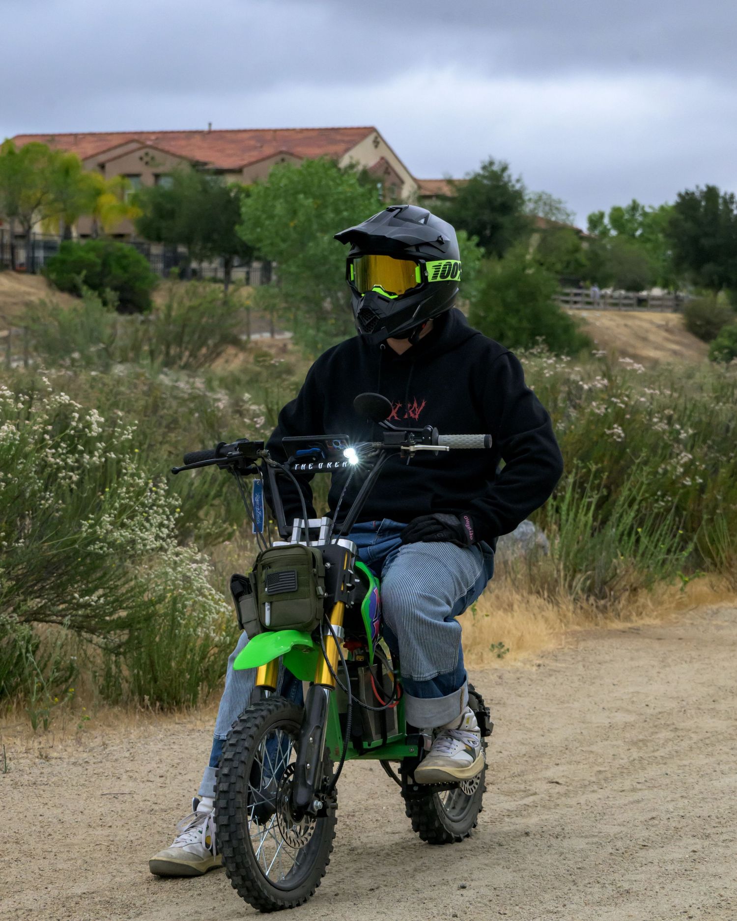 Rider in black hoodie and yellow goggles helmet sits on green dirt bike on dusty trail with overcast sky.