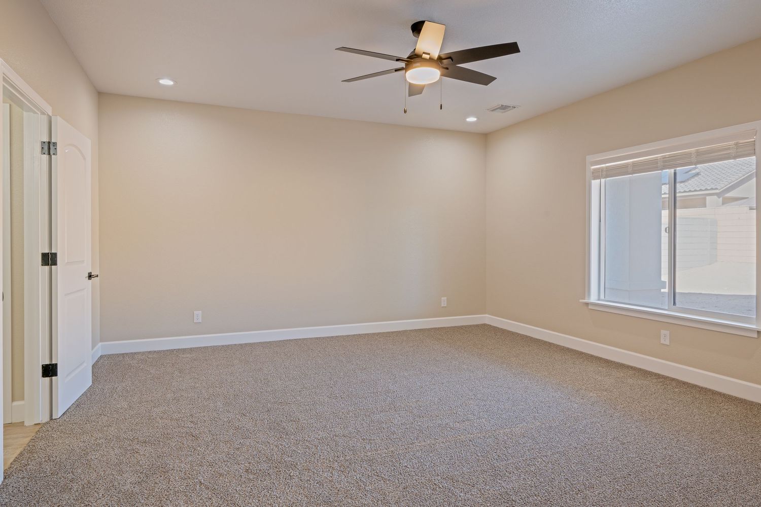 Large empty beige room with recessed lighting, ceiling fan, and window showing potential as a living or family space.