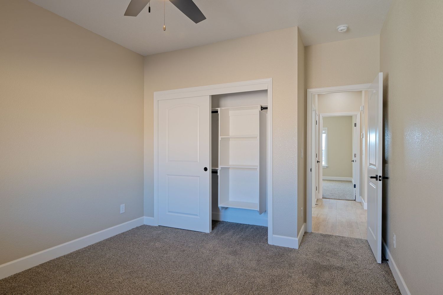 Empty bedroom with beige walls, white doors, ceiling fan, and neutral carpet showing a closet and hallway entrance.