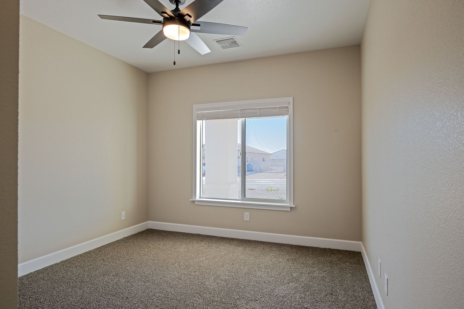 Empty beige bedroom with carpet flooring, ceiling fan, and window showing natural light.