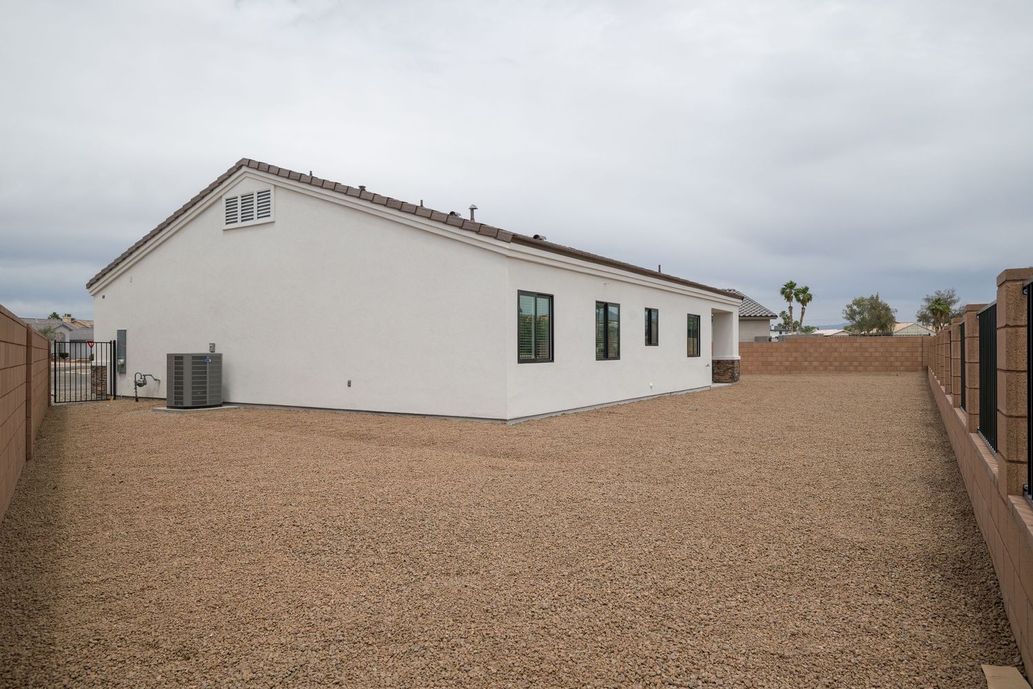 White stucco home exterior with gravel backyard, green windows, AC unit, and block wall fencing under cloudy skies.