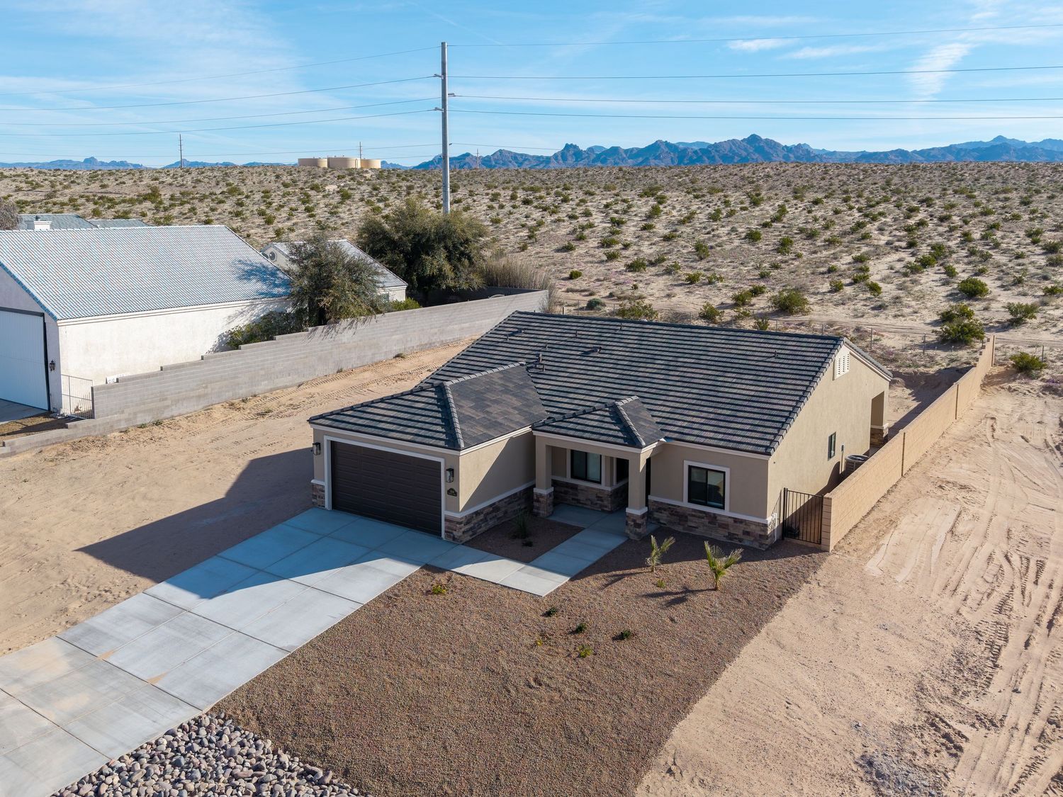 Aerial view of a single-story desert home with black roof and concrete driveway surrounded by arid landscape and mountains.